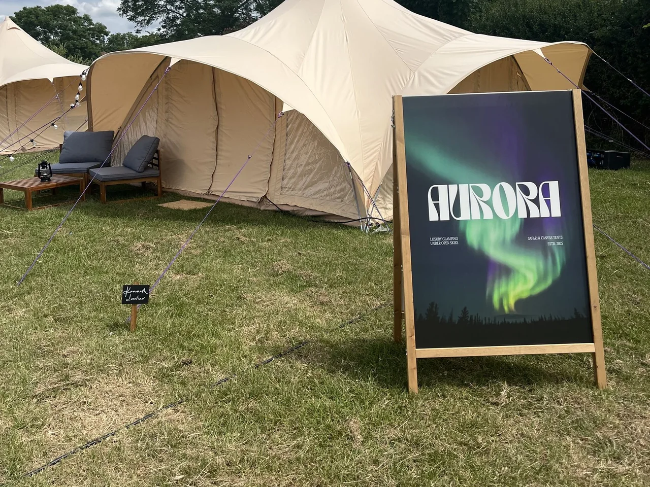 An Aurora A-board with a green aurora printed on it, standing in grass beside a cream canvas canopy