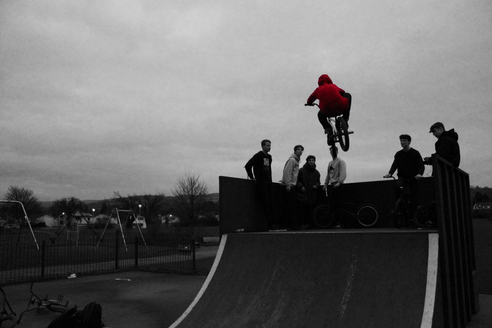 A BMX rider in a red hoodie airborne above a skatepark ramp, everything else in black and white, with the rest of the crew watching from the deck