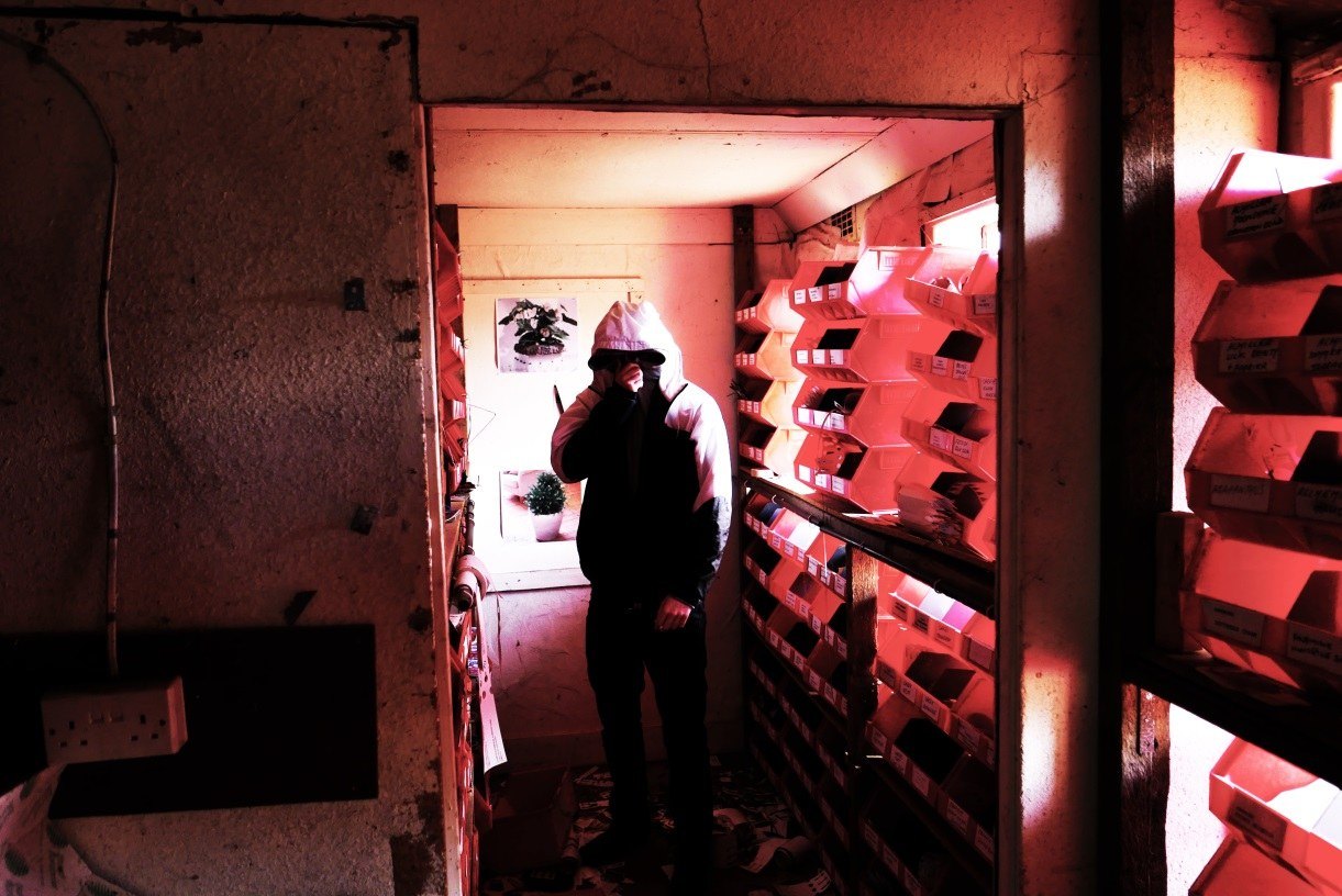 A narrow corridor lined with red racking, lit entirely red, with a figure standing at the far end silhouetted against a bright doorway