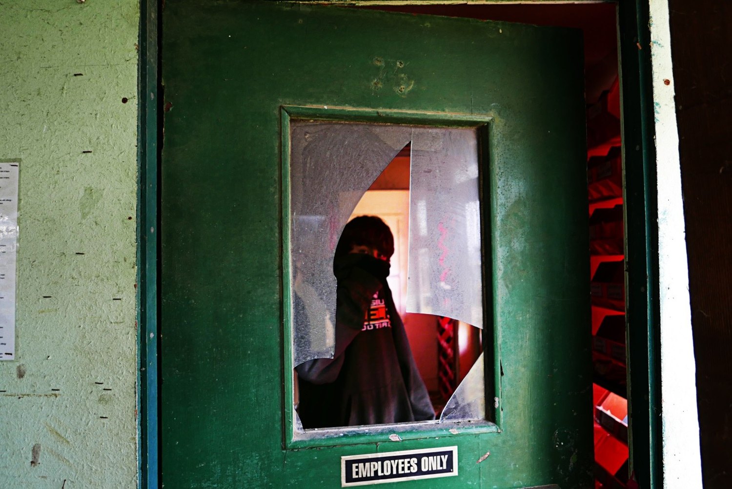 A green painted door frame with a smashed window, an Employees Only sign, and a figure in a hood visible through the broken glass in a red-lit room beyond