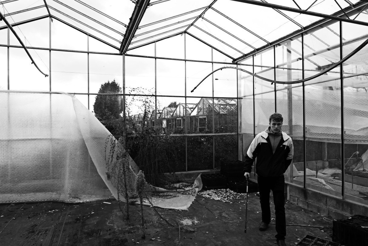 Inside a derelict greenhouse, torn polythene hanging from the roof frame, a lone figure standing among the debris