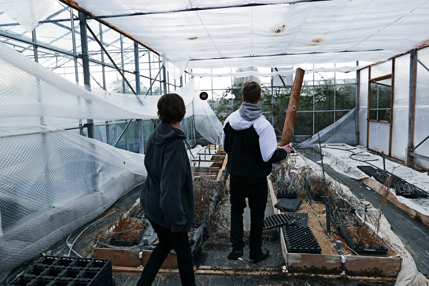 Two figures standing on raised beds inside a long abandoned greenhouse, the roof structure receding to a bright far wall