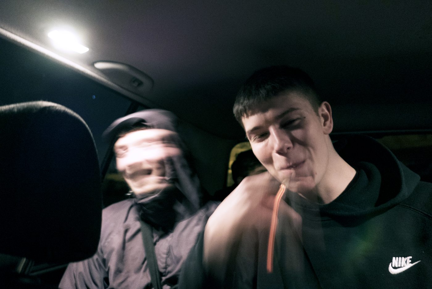A portrait taken in a car at night, interior light on the subject's face, a second figure blurred by motion beside him