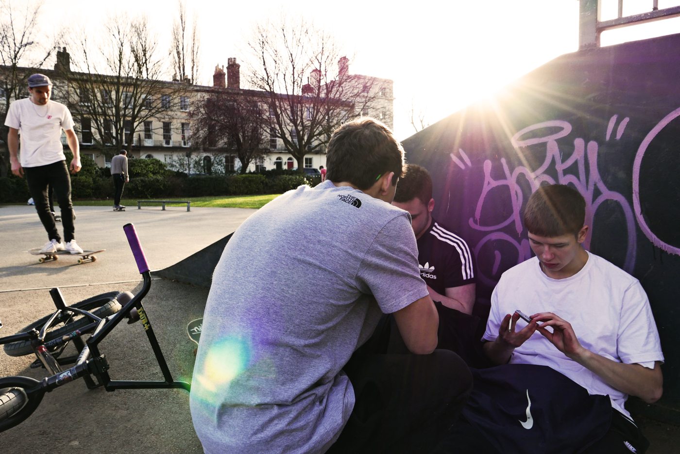 Two young men sitting on a skatepark ramp at sunset, backlit, with a BMX laid down beside them and a third rider in the background