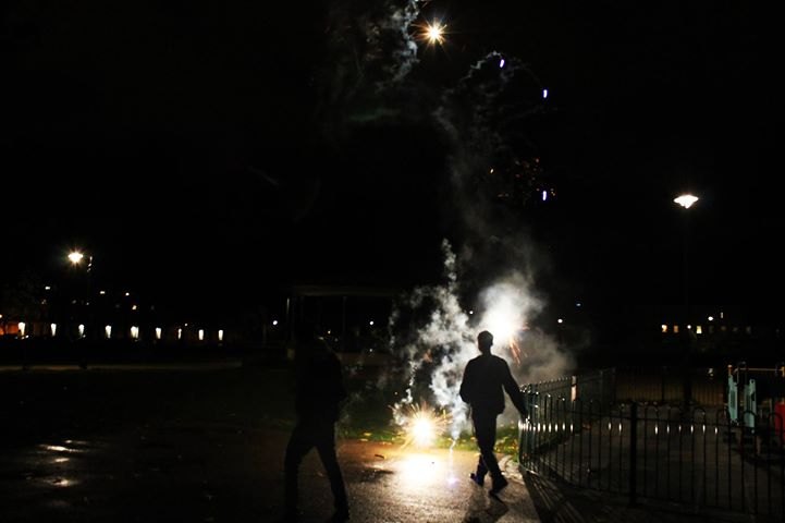 Two figures silhouetted against a firework going off at ground level in a dark park, street lights and railings behind them