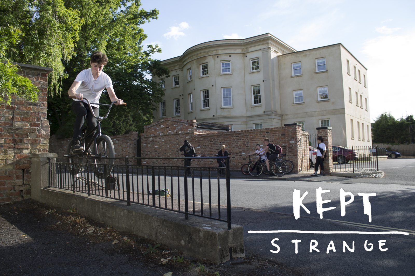 A rider on a BMX balanced on a stone wall outside a Georgian building, with the hand-lettered Kept Strange wordmark set into the lower right of the frame