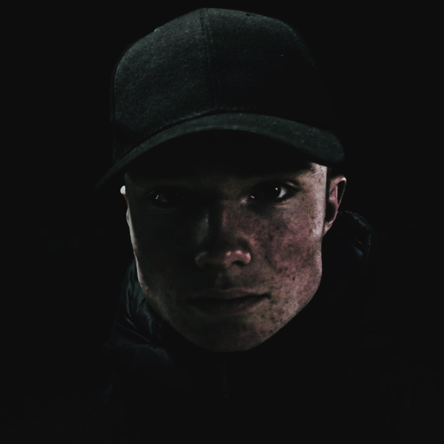 A low-key portrait of a young man in a flat cap, the brim throwing his eyes into shadow