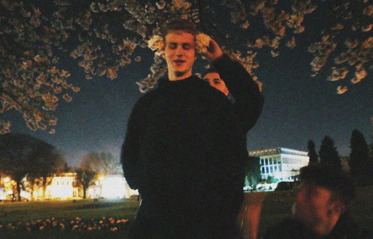 A night portrait: a young man standing under a cherry tree in blossom, the lights of a town spread out behind him