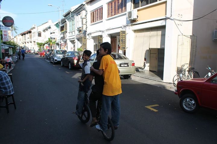 A street in George Town, Penang: low shophouses, parked cars, a group of men talking in the road