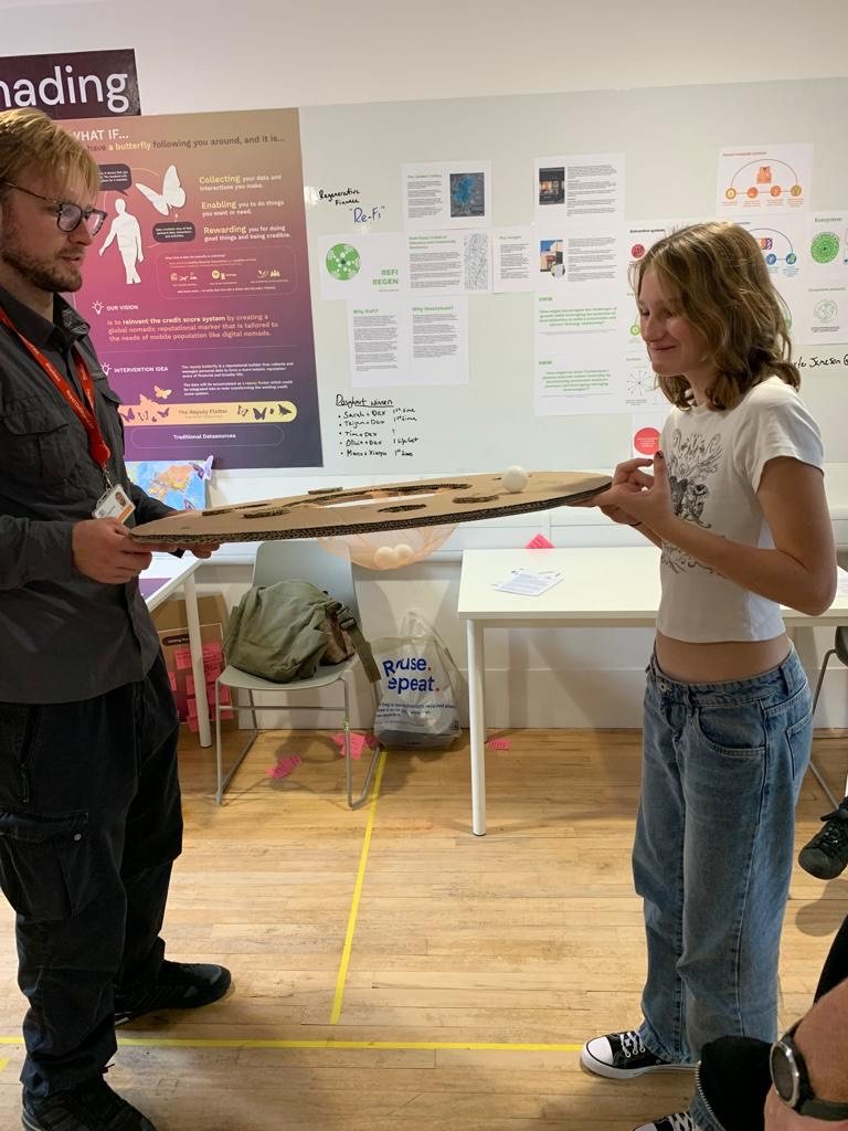 Two people at an exhibition stand holding opposite edges of a large green and red cardboard doughnut between their fingertips, trying to keep ping pong balls balanced on it