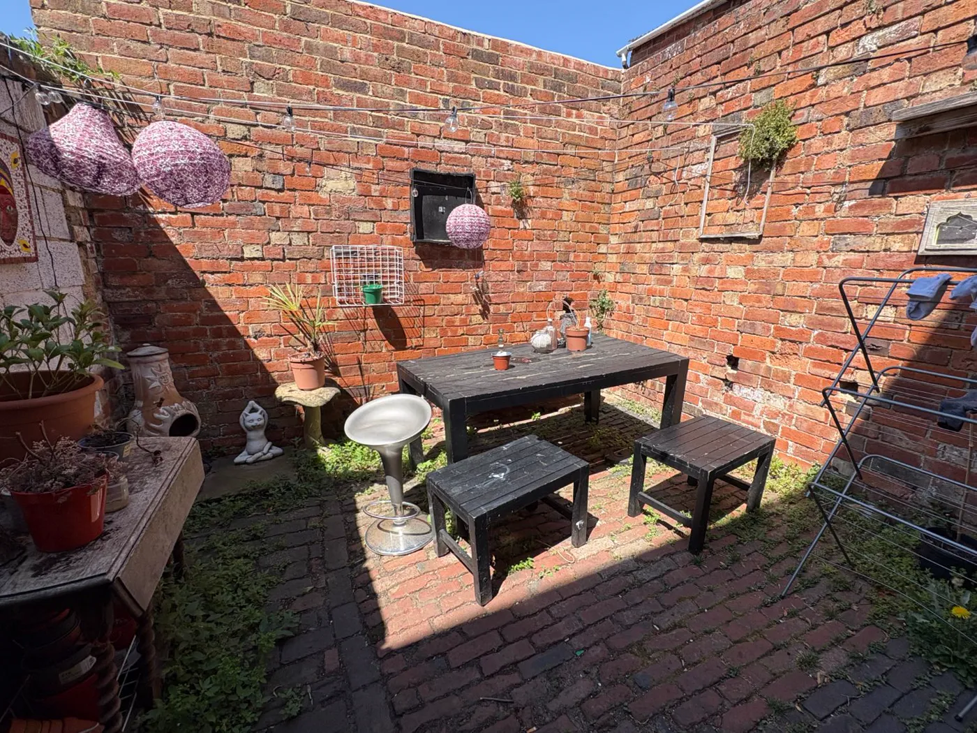 Photograph of a small walled back yard: red brick, a weathered picnic table, paper lanterns and potted plants