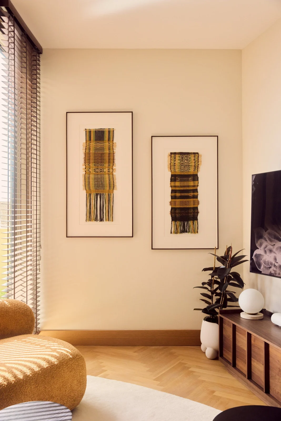 Two framed woven textile pieces on a pale wall, above herringbone oak and an ochre chair
