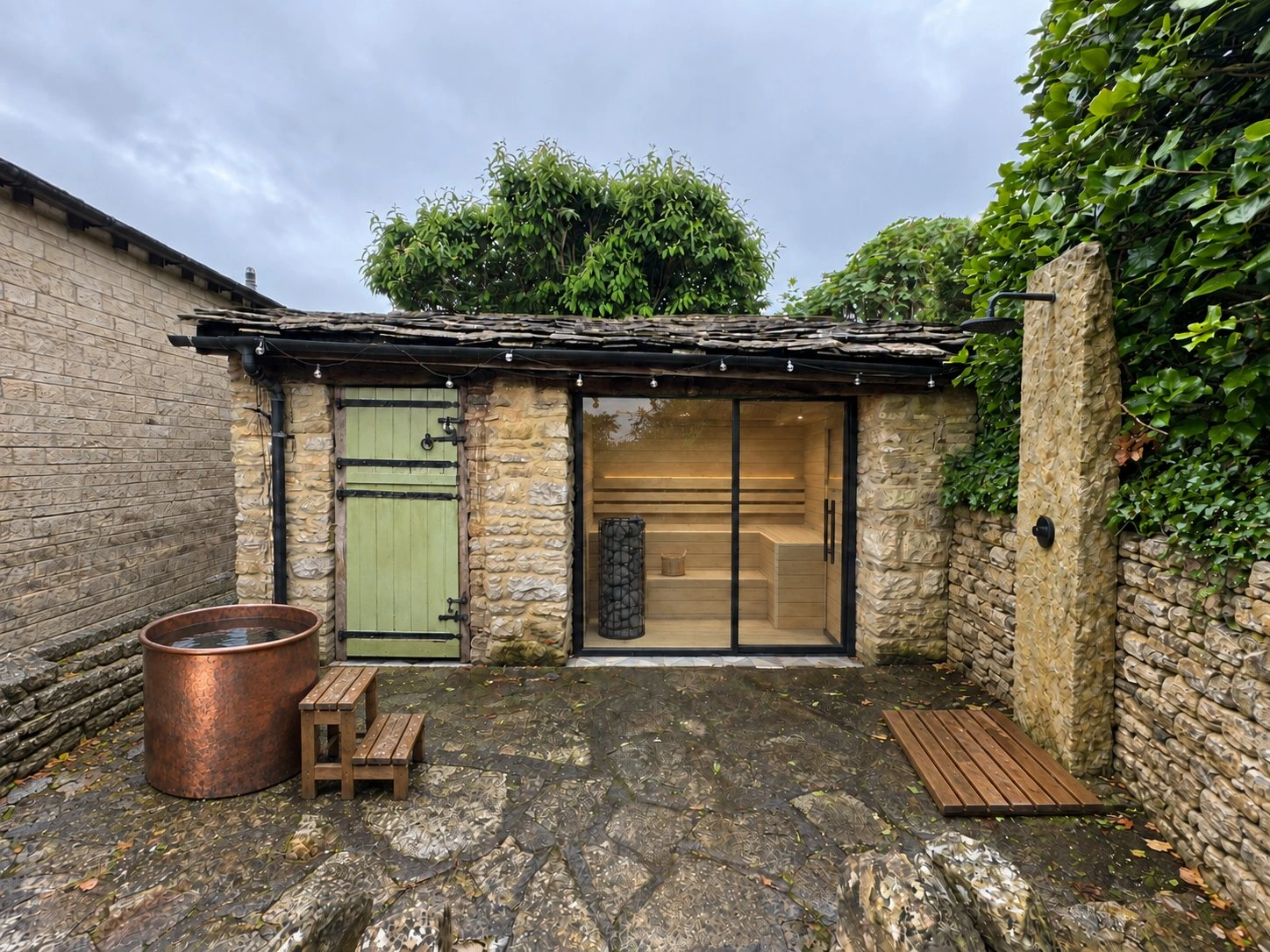 The same yard from the same camera with the design in it: a glazed sauna in the outbuilding, a copper plunge tub, a slab shower and a teak duckboard.
